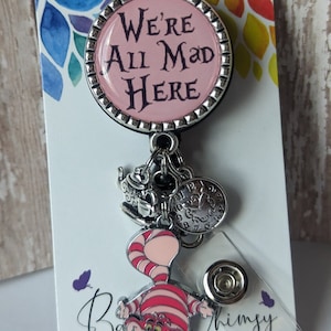May include: A pink and silver badge reel with a Cheshire Cat charm and a teacup charm. The badge reel has a pink circle with the text "We're All Mad Here".