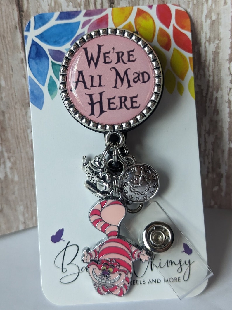 May include: A pink and silver badge reel with a Cheshire Cat charm and a teacup charm. The badge reel has a pink circle with the text "We're All Mad Here".