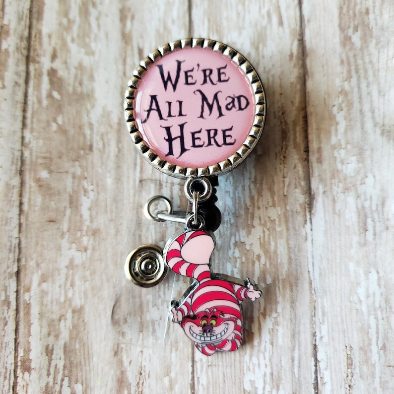 May include: A pink and silver retractable badge holder with a Cheshire Cat charm. The badge holder features a round, silver-rimmed button with the text "We're All Mad Here".
