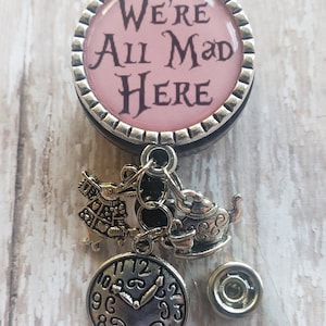 May include: A pink and silver badge holder with a quote from Alice in Wonderland, "We're All Mad Here". The badge holder has a silver charm of a teapot, a clock, and a character from Alice in Wonderland.