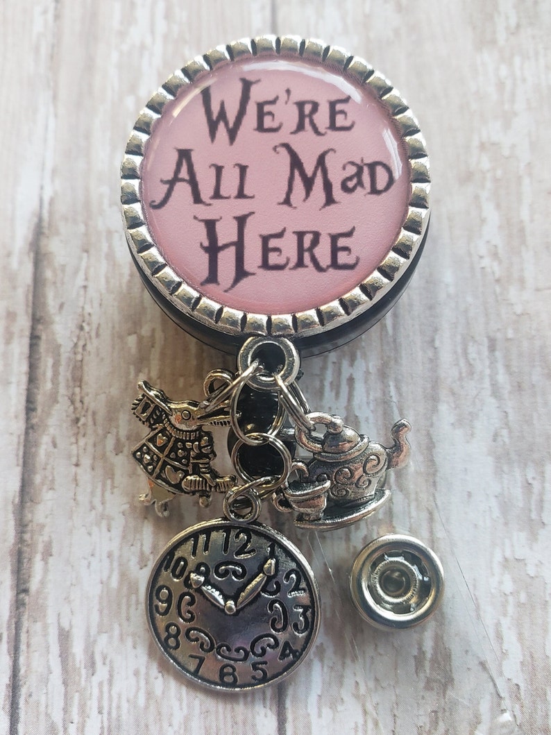 May include: A pink and silver badge holder with a quote from Alice in Wonderland, "We're All Mad Here". The badge holder has a silver charm of a teapot, a clock, and a character from Alice in Wonderland.