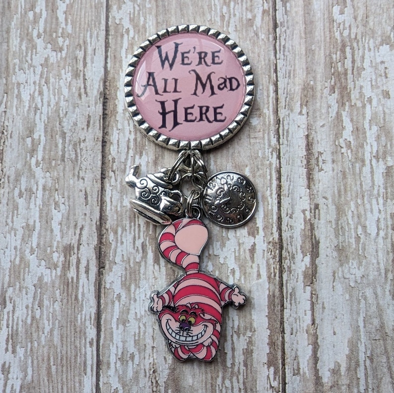 May include: A silver charm bracelet with a pink round charm that says "We're All Mad Here" and other charms including a Cheshire Cat, a teapot, and a clock.