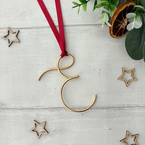 May include: A gold-coloured metal letter "E" Christmas ornament with a red ribbon. The ornament is surrounded by small wooden star cutouts and greenery. The background is a white wooden surface.