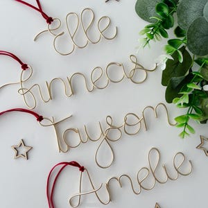 May include: Four gold wire ornaments with the names "Elle", "Finneas", "Taysom", and "Amelie" spelled out. Each ornament has a red ribbon attached.