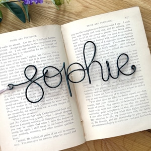 May include: A black metal wire sculpture of the name "Sophie" is displayed on top of an open book. The book is titled "Pride and Prejudice" and the text on the page is visible.