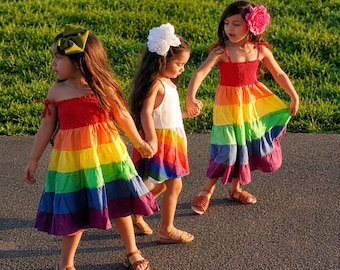 May include: Three children wearing tiered dresses with rainbow-coloured horizontal stripes. One child wears a solid white dress with a rainbow hem, while the others wear full rainbow dresses. They have large hair accessories in olive, white, and pink.
