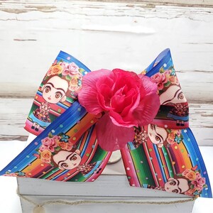 May include: A large blue and red bow with a pink flower in the center. The bow is decorated with a pattern of colorful stripes and polka dots. The bow features a repeated image of a cartoon Frida Kahlo.