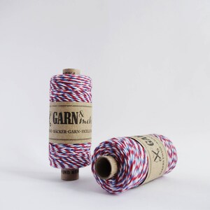 May include: Two spools of red, white, and blue baker's twine. The twine is wrapped around cardboard spools with brown paper labels that read "GARN &  inche" and "GARN".