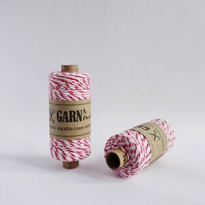 May include: Two spools of red and white baker's twine. The twine is wrapped around cardboard spools with a brown paper label that reads "GARN & more" and "TWINE - B&Auml;CKER - GARN - FICE"