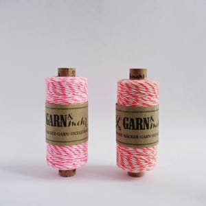 Könnte beinhalten: Zwei Spulen pinkfarbener und weißer Bäckergarn mit braunen Papierretikeln, auf denen "GARN & mehr" und "WINE-BACKER-GARN-FICELLER" steht.