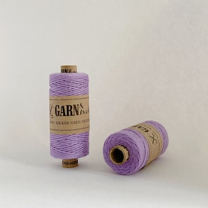 May include: Two spools of lavender coloured twine, each with a brown paper label that reads "GARN & mehr" with a pair of scissors icon. The label also includes the text "TWINE - BÄCKER-GARN - FICELLI - CORD - MEHR"