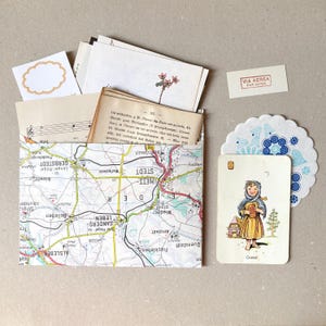 Op de afbeelding: Een papieren envelop gemaakt van een landkaart met Duitse plaatsnamen, gevuld met vintage papier, bladmuziek en een bloemenillustratie. Daarnaast liggen een kaartje met een kind genaamd Gretel, een blauw patroon onderzettertje en een Via Aerea-label.