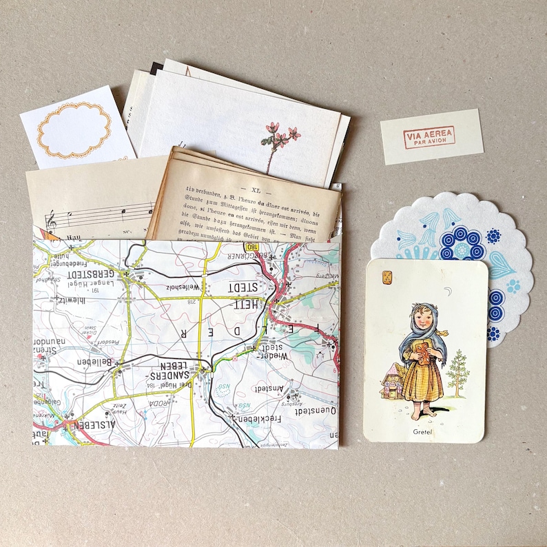 Op de afbeelding: Een papieren envelop gemaakt van een landkaart met Duitse plaatsnamen, gevuld met vintage papier, bladmuziek en een bloemenillustratie. Daarnaast liggen een kaartje met een kind genaamd Gretel, een blauw patroon onderzettertje en een Via Aerea-label.