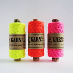 Könnte beinhalten: Drei Spulen Neon-farbener Schnur. Die Spulen sind mit "GARN &" und "TWINE - BÄCKER - GARN - FICELLI" beschriftet. Die Farben sind Neon-Gelb, Neon-Orange und Neon-Pink.