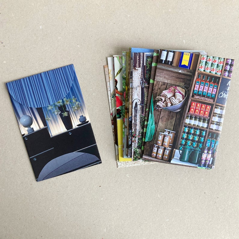 May include: A stack of colorful postcards with various images, including a blue and white striped design, a floral pattern, and a scene of a grocery store with shelves stocked with canned goods.