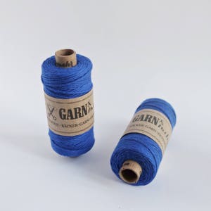 Könnte beinhalten: Zwei Spulen blauer Paketschnur mit braunen Papierretikeln, auf denen "GARN & TWINE - BÄCKER-GARN - FICH...