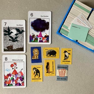 Op de afbeelding: Een verzameling spelkaarten en kleine rechthoekige papieren labels. Drie kaarten tonen de nummers 6, 7 en 8 met illustraties van weer- en stadstaferelen. Verschillende kleinere labels tonen dieren zoals een leeuw, beer, pingu&iuml;n en chimpansee.