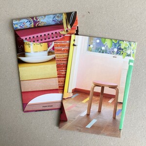 May include: Two folded paperbacks with colorful collage covers. The left cover features a teacup and saucer on a stack of books with text from the book covers. The right cover features a pink and white room with a wooden stool and a green and white patterned border.