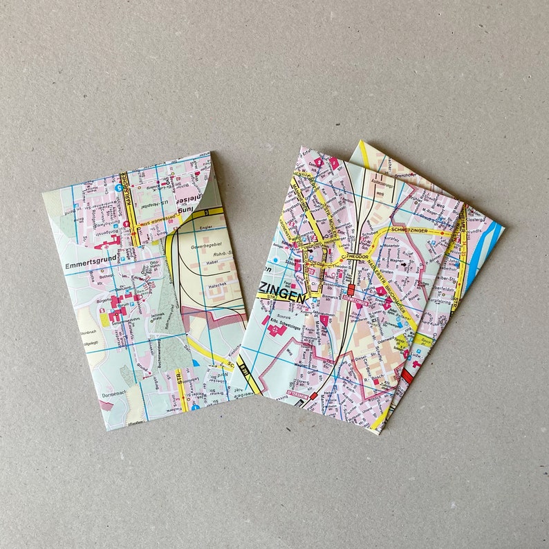 May include: Three folded paper envelopes with a map of a city printed on them. The map is in shades of pink, yellow, and blue. The text on the map includes the words "Emmertsgrund" and "Zingen".