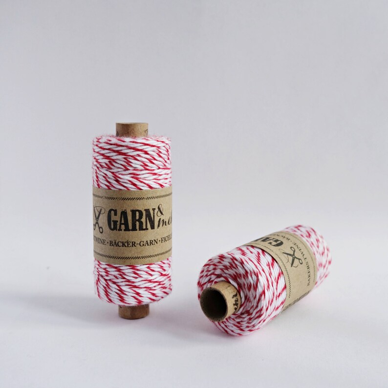 May include: Two spools of red and white baker's twine. The twine is wrapped around cardboard spools with a brown paper label that reads "GARN & more" and "TWINE - B&Auml;CKER - GARN - FICE"