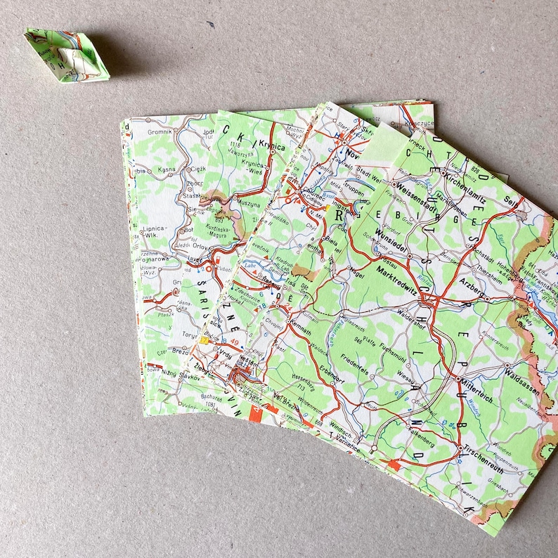 Op de afbeelding: Een klein origamibootje gevouwen van een landkaart ligt naast een stapel vierkante papieren servetten met een gedetailleerde kaart van de regio rond Wunsiedel, Marktredwitz en de Tsjechische grens, met groene, witte en rode wegmarkeringen.