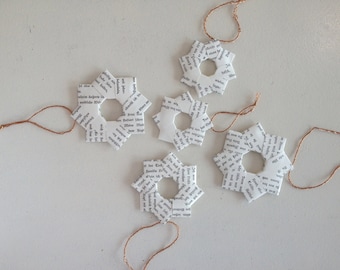 May include: Five white paper snowflake ornaments with brown twine hanging from each. The snowflakes are made from pages of a book and have black text printed on them.