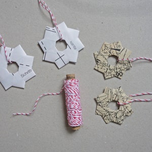 May include: Four paper star ornaments with red and white twine attached. The ornaments are made from paper with different patterns, including a map and a pattern with the words "Schritte" and "Kante". A spool of red and white twine is in the center of the image.