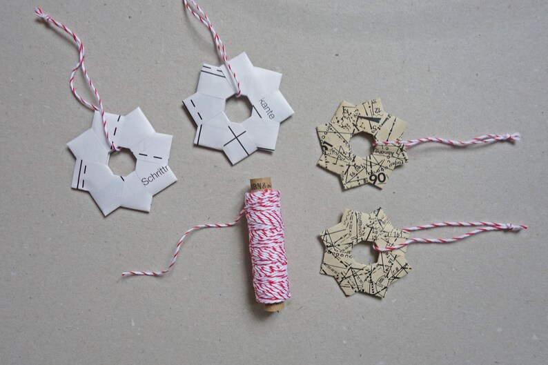 May include: Four paper star ornaments with red and white twine attached. The ornaments are made from paper with different patterns, including a map and a pattern with the words "Schritte" and "Kante". A spool of red and white twine is in the center of the image.
