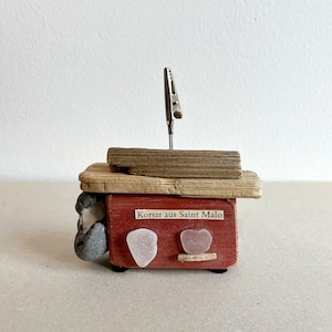 Può includere: Un piccolo modello di barca in legno rosso con un ponte in legno consumato e una clip di metallo in cima. La barca ha un cartello che recita "Korsar aus Saint Malo" e due pezzi di vetro di mare bianco sul lato.
