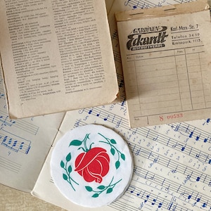 Op de afbeelding: Een verzameling vintage papieren voorwerpen, waaronder een woordenboekpagina, een beige ontvangstbewijsblok met de tekst Gardinen Eckardt Karl-Marx-Str. 7, een blad met muzieknotatie en een ronde witte onderzetter met een rode roos en groene bladeren.