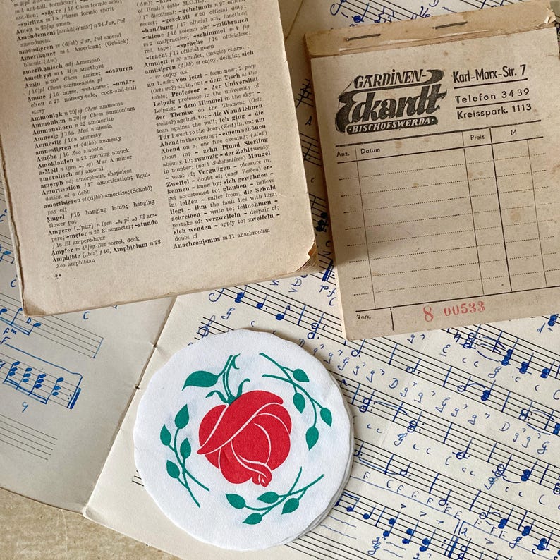 Op de afbeelding: Een verzameling vintage papieren voorwerpen, waaronder een woordenboekpagina, een beige ontvangstbewijsblok met de tekst Gardinen Eckardt Karl-Marx-Str. 7, een blad met muzieknotatie en een ronde witte onderzetter met een rode roos en groene bladeren.