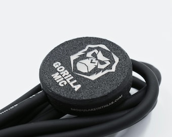 May include: A black circular microphone component with a textured surface featuring a silver gorilla head logo and the text GORILLA MIC. The device is attached to a thick black cable with the text modularsynthlab.com printed on the side edge.