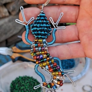 Pode incluir: Um chaveiro de lagarto de contas com um porta-chaves prateado. O lagarto tem uma cabeça e cauda de contas azuis, com um corpo de contas multicolorido. As pernas e os braços são feitos de arame de prata. O chaveiro é segurado em uma mão.