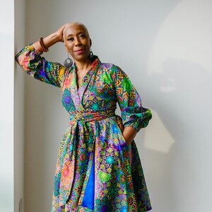 May include: A woman wearing a colorful wrap dress with a floral pattern. The dress has a blue sash and a long, flowing skirt. She is wearing pink heels.