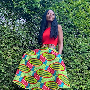 May include: A woman wearing a red sleeveless top with a brown belt and a green, red and blue patterned skirt. She is standing on a wooden deck with a green bush in the background.
