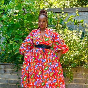 May include: A woman wearing a red, orange, green, and blue paisley print dress with a black belt. She is wearing pink high heels.