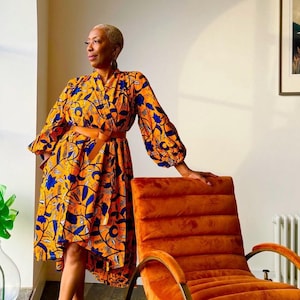 May include: A woman wearing a yellow and blue patterned dress with a brown belt. She is standing next to an orange velvet armchair with gold metal legs.