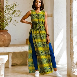May include: A sleeveless, floor-length tunic with a yellow and blue patterned design. The tunic has a high slit on each side and is paired with blue jeans and white sneakers. The model is wearing gold hoop earrings and a watch.