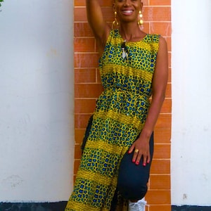May include: A woman wearing a yellow and blue patterned dress with a side slit. She is standing in front of a brick wall and has her hand raised above her head.