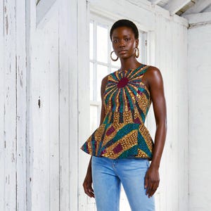May include: A sleeveless, patterned top with a peplum design. The top features a vibrant mix of colors, including gold, teal, and burgundy, arranged in a sunburst pattern. The model is wearing light blue jeans. The background is a white-painted room with a window.