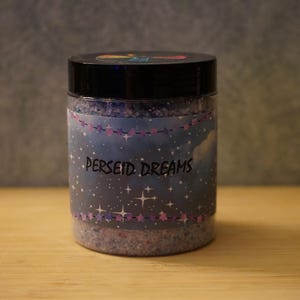 Peut inclure: Un pot en plastique transparent avec un couvercle noir, rempli d'une substance granulaire. Le pot porte une étiquette avec les mots "PERSEID DREAMS" en blanc, ainsi qu'un motif de ciel étoilé. Le pot est posé sur une surface en bois.