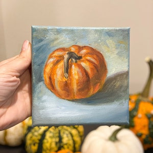 May include: A small painting of a single orange pumpkin on a blue background. The pumpkin has a dark brown stem and is slightly tilted to the right. The painting is on a canvas and is framed.
