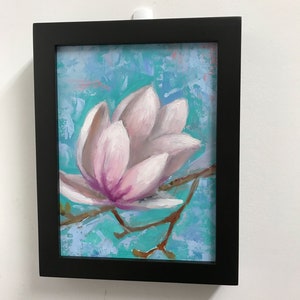 May include: A framed painting of a pink magnolia flower with a light blue and white background.