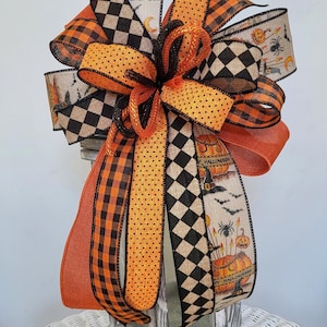 May include: A decorative Halloween bow featuring orange, black, and cream ribbons. The bow includes polka dots, checkered patterns, and ribbons printed with pumpkins, bats, and the words "Happy Halloween".