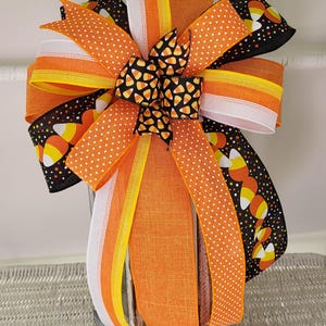 May include: A decorative bow featuring orange, white, yellow, and black ribbons. The bow has a central knot with candy corn pattern, and the ribbons are adorned with polka dots and candy corn designs. Perfect for Halloween decorations.