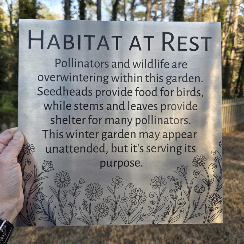 May include: A rectangular metal garden sign with the words "HABITAT AT REST" in black. The sign has a floral border and text describing the garden's purpose for pollinators and wildlife during winter.