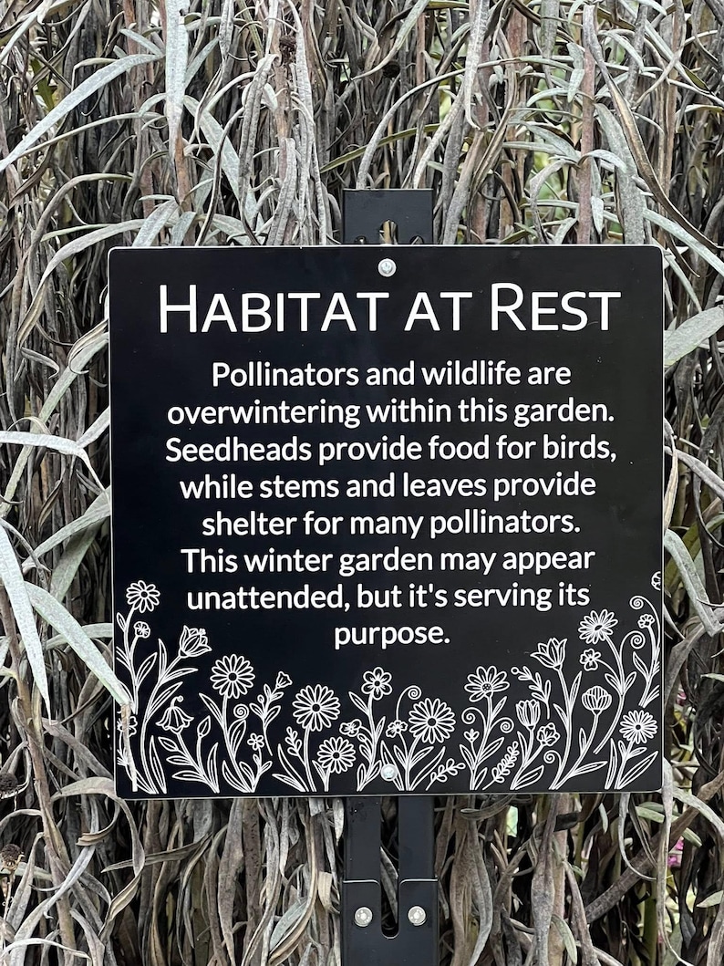 May include: A black rectangular sign with white text that reads "HABITAT AT REST." The sign explains the garden's purpose for pollinators and wildlife during winter. Floral illustrations decorate the bottom edge of the sign.