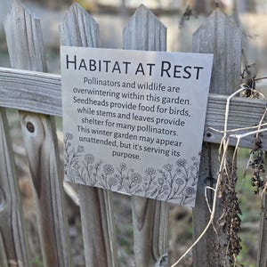 May include: A silver metal sign with the words "HABITAT AT REST" in bold, black letters is attached to a wooden fence. The sign's text describes the importance of a winter garden for pollinators and wildlife. A floral border decorates the bottom of the sign.