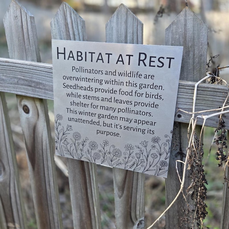 May include: A silver metal sign with the words "HABITAT AT REST" in bold, black letters is attached to a wooden fence. The sign's text describes the importance of a winter garden for pollinators and wildlife. A floral border decorates the bottom of the sign.