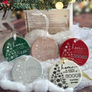 May include: Five round ceramic ornaments with a snowflake design and the text "home is where your heart is" with a city and state name and zip code. The ornaments are in various colors including green, pink, red, blue, and white.
